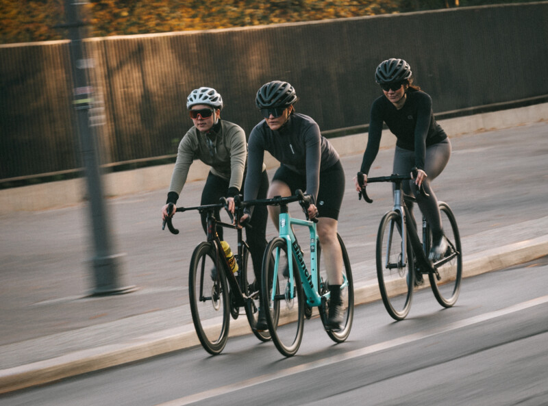Eine Frau lächelt in die Kamera, trägt ein schwarzes T-Shirt mit dem Logo von "Deutsche Dienstrad" und steht auf einer Terrasse mit Blick auf Gebäude und Bäume im Hintergrund.