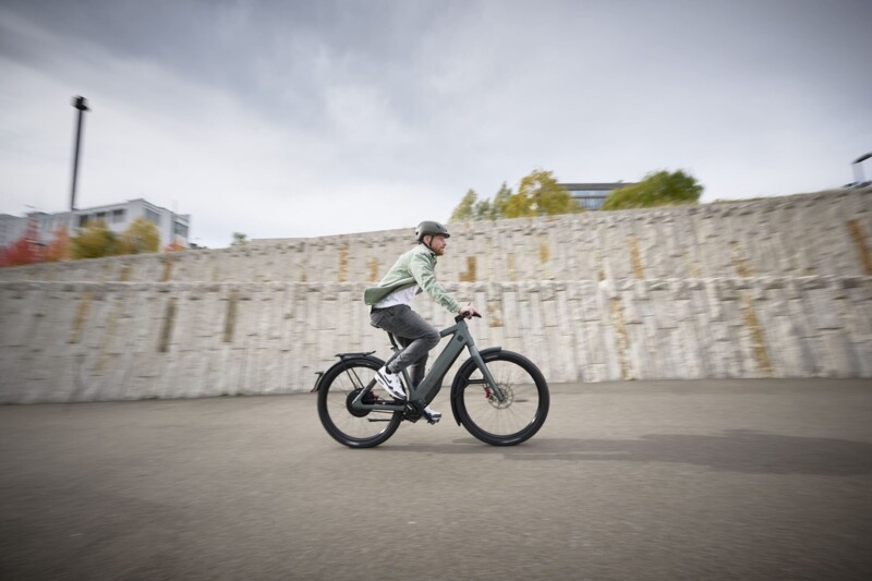 Ein Mann mit Helm fährt auf einem E-Bike vor einer Steinmauer, während der Himmel wolkenverhangen ist.